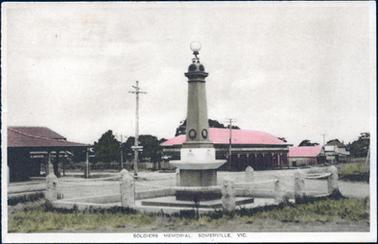 This image shows a Soldiers Memorial in the foreground surrounded by concrete posts connected by chains.  There are shops across the road with red roofs and verandahs over the footpath.  The shop on the corner, on the left-hand side of the image has a sign ‘S. Lacer’ printed on it.  Electricity poles and street signs can be seen and there is grass in the foreground.  Large trees  can be seen across the road behind a fence.  Printed at the bottom of the image is: “Soldiers Memorial, Somerville. Vic.”. 