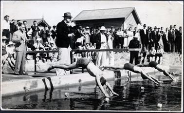 This image shows three people diving into a swimming pool.  A large crowd of well-dressed people are looking on. Three officials are standing near the edge.  A low diving board is at the centre of the image.  Two small buildings are behind the onlookers.  