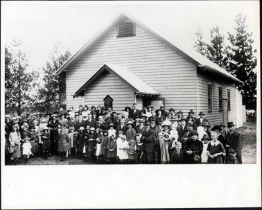 This image shows a large group of over one hundred men, woman and children standing outside a weather-board building, posing for a photo.  They are standing in several rows with most of the children in the front row.  Everyone is very well dressed with the majority wearing coats.  The building behind them has a narthex at the front with a small side opened door and  a sign on the front which reads: “Baxter Public Hall”.  An air vent is seen at the top of the front wall, near the roof line.  Three windows and a small door are along the side of the building. Tall pine trees are on either side of the image.  Part of a harnessed horse can be seen on the far right-hand side beside a partly visible water tank.  