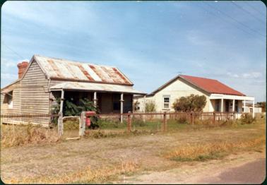 This image shows two older style houses.  The older house on the left of the photograph is weatherboard and not painted.  The galvanised iron roof has faded red stripes across it and is rusty in parts.  There is a bull-nosed verandah at the front with four posts.  A large chimney can be seen at the back.  There is a post and wire fence across the front with a little gate and a red letterbox. A picket fence separates the two houses.  The house on the right of the photograph is weatherboard and  painted cream with a red roof.  It has a front verandah with four posts. The grass between the houses and the street looks dead.  The sky is blue with a little high-level cloud.  