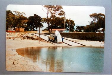 This image shows two people unloading a boat from a four-wheeled drive vehicle which is backed down a boat ramp.  Water is in the foreground which is surrounded by sand.  A brick wall is on the right-hand side.  A red car is parked on the far left-hand side and two cars are parked behind the brick wall.  Buildings can be seen on the far left-hand side, in the middle and on the far right-hand side of the image.  Tall trees are all along the back.  Several power poles can be seen.  