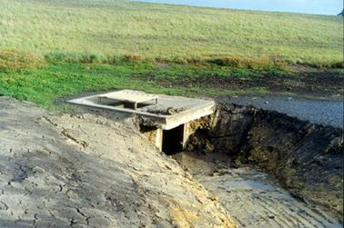 This image shows a deep dirt drain which has been dug in a grassed paddock.  It runs into a square concrete culvert.  There is some muddy water in the bottom of the drain. 