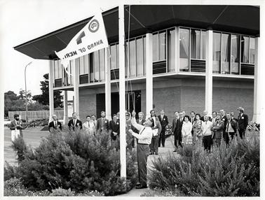 This image shows a large group of approximately thirty-one people standing watching a man raise a white flag.  Printed on the flag is: “Award of Merit”, a partly visible symbol and a map of Australia inside a circle.  The man raising the flag is wearing a long sleeved white shirt, tie, dark trousers and a cap.  Most of the men are wearing suits.  There are only two women in the group.  Shrubs are on either side of the flag pole.  A two story building with white posts is behind the group.  The walls of the top floor of the building are made up of long glass windows.  A tall pole with a light and fencing can be seen to the far left of the image.  