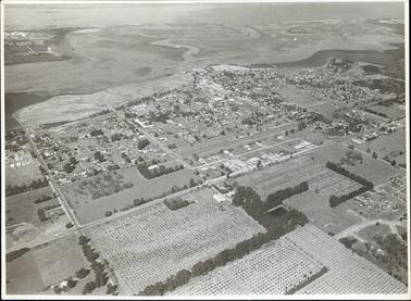 This image is an aerial view of a town.  Rows of threes are at the bottom of the photograph, the town in the centre and the water at the top section.  Oil tanks can be seen at the top left-hand corner of the photograph and a jetty on the far right-hand side.  
