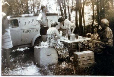 This image shows a group of people enjoying a picnic.  There are four women and two men.  Three of the  women are sitting and they are dressed in summer print dresses.  The fourth woman is standing leaning over the table.  She is wearing a white blouse and print skirt.  The man near the table has no shirt and is wearing swimming trunks.  The other man has no shirt and has a towel wrapped around his waist.  A van with the words “J.A. Babington Hastings Grocery”, is parked behind the group.  Two large wooden boxes are sitting on untidy grass on the ground in the foreground and trees are in the background.  
