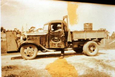 This image shows a truck with two men. One man is sitting in the drivers seat and the other man, who is wearing a white hat, is standing with one leg on the ground and the other in the truck.  His left arm is resting on the open window.  A drum is in the back of the truck.  “Shire of Frankston Hastings 3” is printed on the door.  