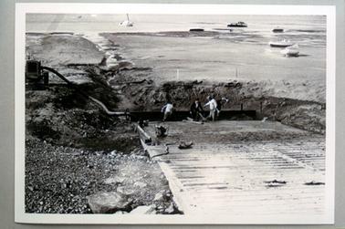 This image shows four men working on a boat ramp with the water and boats in the background.  Some boats are in the water and some on the sand.  A channel has been dug behind the men, running out into the sea.  A machine with a pipe leading down to the workmen is on the bank of the channel.  Gravel and a few large stones are in the foreground on the left-hand side of the image.  