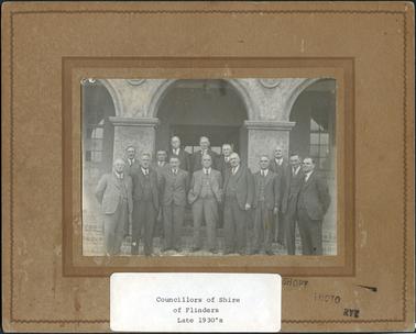 This image shows a group of fourteen men, all dressed in white shirts, ties and three-piece suits standing on the steps outside a building.  The building has two huge pillars and big arches.  Windows can be seen behind the verandah to the left and right of the photograph and a glass door is in the centre.  There are six men in the back row and eight men in the front row.  Most of the men are elderly and the man in the centre, front row, is standing with his hands in his trouser pockets. Typed on white paper and glued on the bottom of the mounting is: “Councillors of Shire of Flinders Late 1930’s”. Also stamped in ink on the mounting is: “Shop Photo Rye”.
