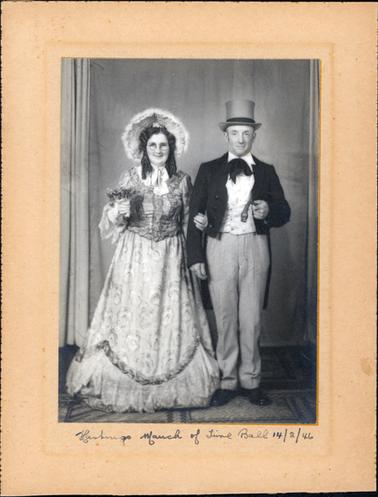 This image shows a smiling woman and man, dressed in old-fashioned ballroom clothes, standing side by side, posing for a photo. The woman is wearing a long sleeved, floor length gown with a large bow at the neck.  The skirt has a pattern of large flowers and has a garland around the bottom.  She is wearing a large bonnet and her hair has ringlets on each side.  She is wearing spectacles and is holding a posy in her right hand.  The man is dressed in top hat, black tie and tails.  He has a white vest, pin-stripped trousers and black shoes.  He is holding a short cane in his left hand.  Curtains can be seen on the wall behind the couple.  Written on the mounting board beneath the photo is: “Hastings March of Time Ball 14/2/46”.