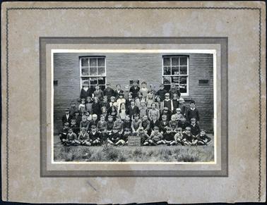This image shows a large group of children, in five rows, posing for a photograph in front of their school.  There are ten children in the back row, fifteen children in the second back row, eleven children in the middle row, fifteen children in the second front row and twelve children sitting on the ground, cross-legged in the front row.  Two boys in the front row are holding a small blackboard which reads: “Hastings School 1098 Grades I to IV”.  The wall behind the children is brick with two large windows.  Three air vents can also be seen along the wall.  