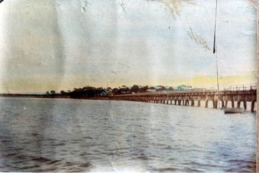 This image shows a long jetty running out into the water with land, buildings and trees in the background.  A small boat is beside the jetty on the far right-hand side.  Brown patches and a black vertical thin line are on the right-hand side of the image.  Small waves run across the water.