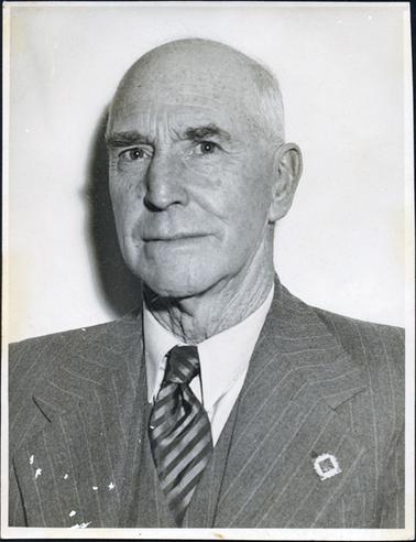 This image shows a portrait of a middle-aged man taken from the chest up.  He is wearing a three-piece suit with a striped tie and white shirt.  He has a badge on his left lapel.  He has thick eyebrows and thinning hair.  