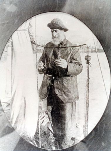 This oval image shows a heavily bearded man standing mending a fishing net.  He is wearing heavy duty coat and pants and hat which is turned up at the front.  Part of a sail and nets and ropes can be seen on the left-hand side of the image.  He is holding net mending tools in each hand.  The background is very light and indistinguishable.  