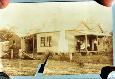 This image shows a small weatherboard cottage with five people on the verandah posing for a photo.  The cottage has a four post verandah across the front, with a set of windows on either side of the front door.  A large chimney and two sets of windows are on the side of the cottage.  A small lean-to is at the back which has small trees and a water tank.  Two more weatherboard buildings can be seen behind the lean-to. There are three women and two young girls on the verandah.  The girls are dressed in white pinafores.  The woman on the left is dressed in a black dress with a white collar.  The other two women, one sitting and one standing, are wearing white blouses and black skirts.  There is a post and wire fence along the side of the property which has a small gate on the left-hand side.  Grass is in the foreground.  Two fingers and two coins are holding the photo down at the four corners.  