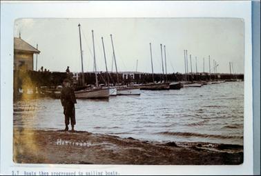 This image shows a  man standing on a foreshore with his left arm stretched out pointing.  He is dressed in dark clothes and wearing a cap.  Behind him can be seen part of a shed, a pier jutting out into the water and a line of fishing boats with tall masts.  Fishing nets are hanging over the railing of the pier.  Several people can be seen along the jetty.  Printed at the bottom is: “2.7 Boats then progressed to sailing boats.”. Also printed in white on the sand is: “Hastings”.