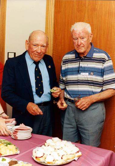 This image shows two elderly men standing behind a table with food in their hands.  They are both looking at the camera.  The man on the left is wearing a dark coat and trousers, blue shirt and tie.  He has a badge on the left lapel of his coat.  His head is bald except for some grey hair on each side.  The man on the right is wearing a blue and green striped polo shirt and green shorts.  He has a name badge on the front of his shirt. The table is set with piles of plates and serviettes and plates of food.  There is a large plate of scones with jam and cream.  Part of a pair of hands can be seen on the left-hand side around a pile of plates.  A brown timber wall is behind the men.  