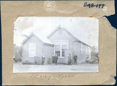 This image is of a wooden building with a small similar building built directly in front.  The large building has two large windows at the front and three sets of windows can be seen along the side.  The front building has one window at the front.  Seven young people are standing outside the buildings.  There are shrubs and trees on either side of the buildings.  A small water tank in up against the front of the building.  Written on the mounting at the bottom is: “S S 3129 Tyabb”.