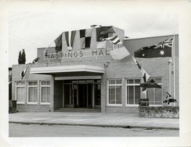 This image shows the frontage of a brick building with the large letters across the top “HASTINGS HALL”.  A covered entrance to the large glass doors is at the centre of the image.  Three large sets of windows are on either side of the entrance.  An Australian flag is flying on a flag pole on the right-hand side of the building.  Flags and bunting are across the front of the building.  A stone fence is on either side of the building.  A footpath and street are in the foreground.  