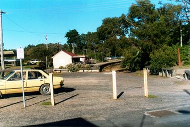 This image shows part of a yellow car and a brown car parked in a car park in front of a railway station.  The station has a red iron roof with a wide undercover verandah at its front.  There is a curved platform and curved rail line which terminates at the car park.  Tall electricity light poles line the platform and cars can be seen parked in the distance.  Power poles stand on the far left-hand side of the image and run along behind the station.  Buildings and another car are on the left-hand side.  White guard posts are in the foreground and a signpost which reads: “V/LINE Car Park Patrons Only”.  Trees and scrubs are in the background and on either side of the rail line.  