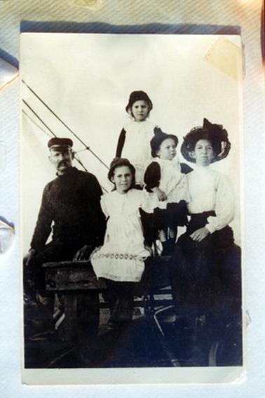 This image shows a family of five posing for a photo.  The man on the left is dressed all in black and is wearing a cap.  He has a moustache.  The woman on the right is wearing a long sleeved, high neck white blouse and a black skirt.  She is wearing a large black hat and spectacles.  The three children are all wearing white sleeveless smocks with black dressed underneath.  The children are all wearing small hats.  