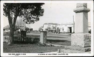 This image shows High Street, Hastings, looking west. Part of the war memorial, with some names visible, is in the foreground with its surrounding wall, and a lady’s bicycle lying on the ground. Behind the memorial is Kraemer’s R.A.C.V. service station, and the hotel is in the background.