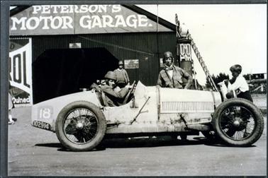 This image shows a racing car parked outside a large shed.  Peterson’s Motor Garage is written in large white letters on the front of the building.  A man is sitting at the wheel, another standing at the side of the car and a small boy is at the front of the car holding a large white jug.  A man is standing at the entrance to the shed.  There is a petrol bowser in front of the garage on the right-hand side of the photo and a ladder is leaning up against the shed nearby.  Part of a petrol sign can be seen on the left-hand side of the photograph.
