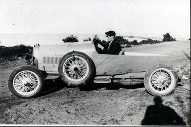This image shows a young man with black hair and dressed in a dark coat, sitting behind the wheel of a racing car.  He is looking straight ahead.  The car has a spare wheel attached to the side.  The shadow of a person is on the foreground  on the right-hand side and water is in the background.  A row of trees and shrubs is between the land and the water and a jetty can be seen jutting out into the water on the far right-hand side.  
