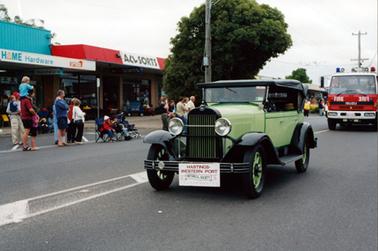 This image shows a green vintage motor car driving down the middle of a street.  It has a black soft top, four doors, green wheel spokes and black mud guards.  Attached to the front is a sign that reads: “Hastings-Western Port Historical Society Incorporated”.  Following behind is a red Isuzu fire truck.  People and shops line the street on the left-hand side of the image.  The two shops the vehicles are passing are: “Home Hardware” and “ACLSORTS”. Two electricity poles can be seen and a large, bushy tree is on the side of the street.  