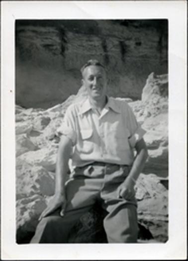 This image shows a man sitting on rocks, posing for a photo.  He is wearing a open-necked shirt with long sleeves rolled up above the elbow.  His hands are resting on his lap and he is smiling.  Rocks can be seen beside him and behind him.  