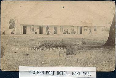 The Western Port Hotel in Hastings. In this image the hotel is a long low single story building with a verandah the full length of the facade. Five women, a man and a child are on the verandah, one woman standing with a bicycle. The foreground of the hotel is open land with some scrubby vegetation and a large tree trunk at the right hand edge. On the image is printed “Wn Port Hotel Hastings”