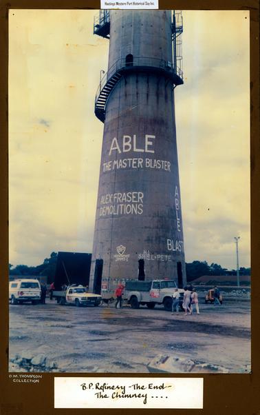 This image shows a tall chimney with steps at the top leading to a doorway.  A landing and a ladder lead on to the top.  Three doorways are at the bottom of the chimney.  Written in large white letters across the lower half of the chimney are: “Able The Master Blaster; Alex Fraser Demolitions; Able Blaster; BP Westernport Jimmy-1; H.I.Shel+Pete”.  A small truck, utility and panel van are parked across the front of the chimney.  People are walking around and a German Shepherd dog is standing between the vehicles.  A tall concrete pole with lights is on the far right-hand side of the image and a high bank with a building on the top is behind the pole.  Trees are in the background and gravel and rock is in the foreground.  