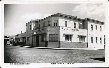 The image is of the Royal Hotel, Hastings.  It is situated on the corner of two streets.  The hotel is on two floors and the name ROYAL HOTEL is clearly visible in large letters on the front and side of the hotel.  There is a car parked on the left-hand side of the hotel in the background.  There are clouds in the sky.  