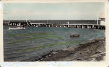 This image shows a jetty stretching out into the water with the foreshore in the foreground.  A man standing in a boat is on the left-hand side of the image and a small row boat is near the shore.  Two people are standing on the jetty.  Tall poles are along the jetty. The printing along the bottom of the post card has been covered in whiteout, however on the copies it reads: “The Rose Series P. 13937 The Jetty, Hastings, Vic.”. 