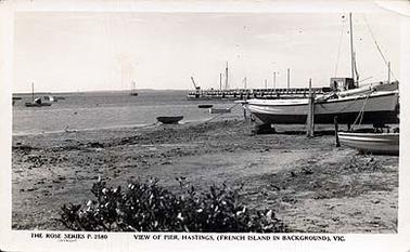 This image shows Hastings pier in the right background. In the right foreground is a fishing boat, moored on dry land, and a dinghy. The foreground shows the mud flats and mangroves, and the background shows Western Port Bay with fishing boats moored and French Island at the horizon.