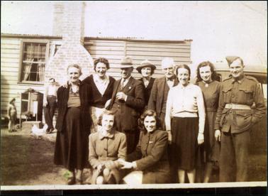 This image shows a group of ten people posing for a photograph.  There are five women and three men standing and two women crouched on the ground in front of them.  They are all wearing winter clothes and smiling.  The man on the right-hand side of the image is wearing an army uniform.  They are standing in front of a motor vehicle.  A weather board house with an iron roof and a large brick chimney is in the background.  A man is leaning against the chimney and a small child is near him.  Between them is a rabbit hutch.  A white chook is on the ground in front of them.  