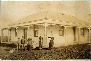 This image shows a cottage with a verandah on two sides.  Three women and three children are standing on the ground in front of the cottage posing for a photograph.  The three women are all wearing dark coloured long dresses.  The little girl has a white pinafore over her dress and pantaloons are showing under her dress.  The two boys are wearing long pants, coats and hats.  A dog is sitting to the left of the boys.  The cottage has four posts on each side of the verandah. Two sets of windows, on either side of the door is on both sides of the house. Rough ground is in the foreground.  Three chimneys can be seen on the roof.   