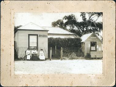 This image shows a weather-board house on the left-hand side of the photograph with the words BOARD & RESIDENCE at the front.  Seated on chairs outside are three women.  The woman on the left of the photograph is patting a dog.  There is a small child standing between the next two women.  The woman on the right has a bouffant hair style and is nursing a cat.  There is a trellis on the wall behind the group.  A large creeper is growing at the front verandah of the house.  A very small section of a water tank can be seen on the left-hand side of the house.  A small fence at the front joins onto a second small weather-board building with the words  POST OFFICE above the window.  On either side of the window are notices stuck to the wall.  A very large tree is behind the Post Office building.  