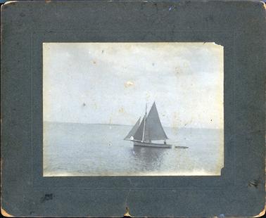 This image is of a boat with sails, towing a tiny boat on a large expanse of water.  Land can be seen on the far horizon and there is a very large expanse of sky.  There are two men on the boat with the sails, one near the tiller and one at the rear of the boat.  