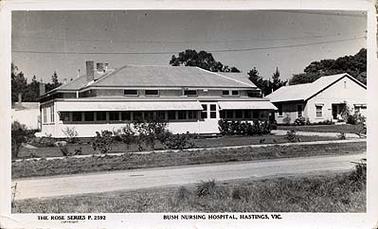 This image shows the Hastings & District Bush Nursing Hospital from the front, a single story brick building with a shaded verandah across the front facade and around part of the right hand side. A smaller building is on the left hand side. There are a footpath and shrubs in front of the building and a gravel road in the foreground.