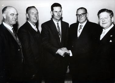 This image shows five men standing, taken from the knees up, posing for a photograph.  All men are dressed in suits, white shirts and ties.  They are all smiling for the camera.  The man on the far right-hand side is shaking hands with the man in the center of the photograph.  