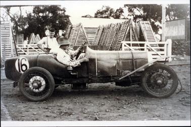 This image shows a man sitting in a racing car.  The number 16 is on the back of the car in large numbers.  Another man is partly seen crouched on the far side of the car and a man is standing watching behind him.  There are many cray coffs in the background.  A white wooden fence and a wooden building with an iron roof can also be seen. Part of a white wooden fence is on the right-hand side of the image.  A large pole is behind the fence.  Large trees surround the property.  