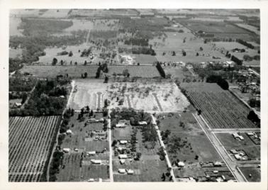 This image shows an aerial view of a small town with paddocks, orchards and roads.  New ground for housing, showing new streets, is in the middle of the image.  