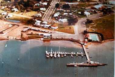 This aerial photo shows a foreshore with a jetty in the foreground and a township above it.  There are two rows of boats anchored on the left-hand side of the jetty and several small boats along the right-hand side of the jetty.  A street leads down to the jetty with a swimming pool on the right.  Several boats are lined up on the land.  Motor vehicles are parked on the foreshore, facing the bay.  Streets, buildings and houses are in the top half of the image.  