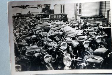 This image shows a large collection of motor bikes parked very close together outside a weather board building.  Two old model motor vehicles are parked beside them.  The building has a galvanized iron rain water tank on a wooden stand against the wall and two sets of windows can be seen along the wall.  A tree branch can be seen in the upper left-hand side and other trees in the distance.  