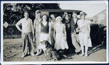 This image shows a group of eight people standing in front of a motor vehicle, posing for a photograph.  A dog is sitting in front of the group looking to the left-hand side of the photograph.  There are three men and five women.  Everyone is smiling.  The young woman at the front on the right-hand side is wearing a hat and holding a tennis racket in her right hand.  The other two young women at the front are wearing sleeveless vests and berets.  The two women standing behind are older and the three men are wearing coats and jumpers.  Part of a building can be seen on the right-hand side of the image and another building is behind a fence and trees on the left-hand side of the image.  A sign displaying an emblem is attached to the fence.  