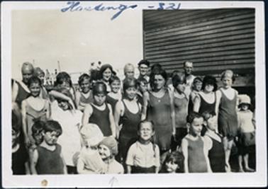 This image shows a large group of approximately twenty-five children and three adults posing for a photo beside a weather-board building.  They are all dressed in swimming attire.  The three adults, two men and one woman are standing in the back row.  Part of a jetty with people and boat masts can be seen in the background.  Written in ink at the top of the photograph is: “Hastings - 32”.