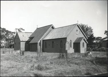 This image shows a church consisting of three buildings joined together.  There is a weatherboard  building at the front with an iron roof and a narthex at the front.  Behind this building is a brick building with a tile roof and older wooden building with an iron roof is at the rear. A cross is at the top of the front building and another cross can be seen at the back of the middle building.  Small trees are seen in the foreground surrounded by stakes and wire netting.  Untidy grass is all around the building and there are large trees in the background.  Part of a  building can be seen off to the right of the photo.  There is a noticeboard in the foreground.  