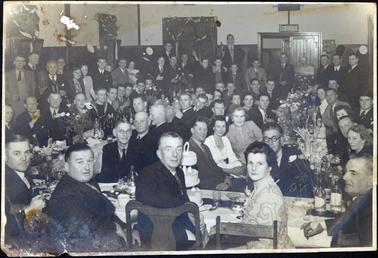 This image shows a large group of approximately seventy people in a hall, enjoying a celebration.  The large crowd are sitting at three long tables covered in decorations, bottles and glasses.  A large three-tier, iced cake is on the top table.  Many other men and women are standing.  There are decorations on the walls of the hall and a door with an exit sign is at the back of the hall.  Everyone is very well dressed.  A man, standing on the left-hand side of the image is holding a football.  