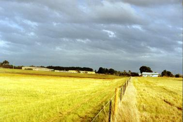 This image shows a large grassed paddock in the foreground with an airfield on the left-hand side in the distance.  A post and wire fence is running down the centre of the image.  A building can be seen on the right of the fence and sheds and small aircraft can be seen on the left-hand side of the fence.  Tall trees are in the background and grey clouds above.  