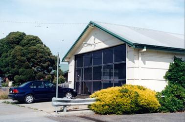 This image shows the front and part of a side wall of a white coloured building.  It has a galvanised iron roof and green painted guttering.  A large glass-panelled door is across the front of the building with a triangular piece of wall above it. A light is over the door.  A dark blue coloured sedan motor car is parked at the front of the building.  A steel post and guard rail is in the foreground.  A large tree is behind a wooden fence on the left-hand side of the image.  Part of a green roof can be seen behind the tree.  A yellow shrub and a green creeper are in the foreground.  